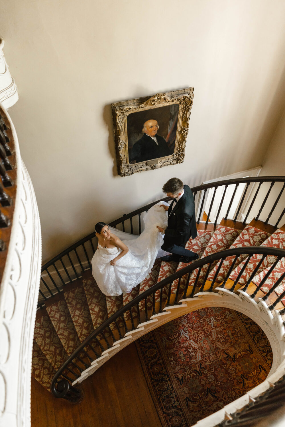 groom holding brides dress as they walk down grand staircase together