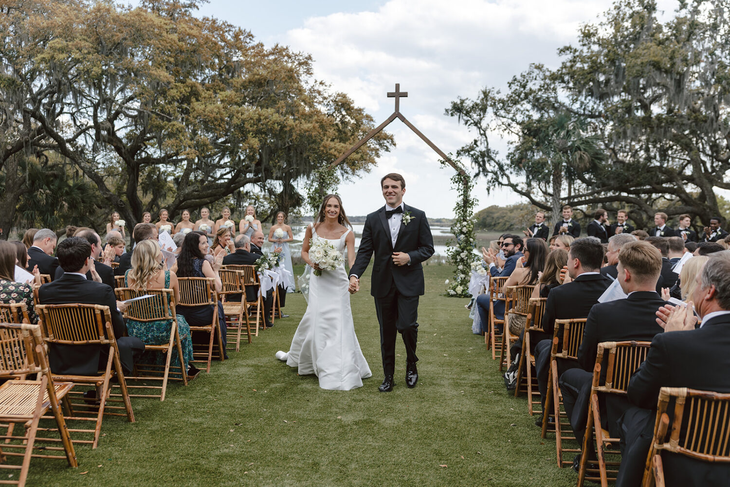 smiling bride and groom walk hand in hand down wedding aisle on grassy lawn